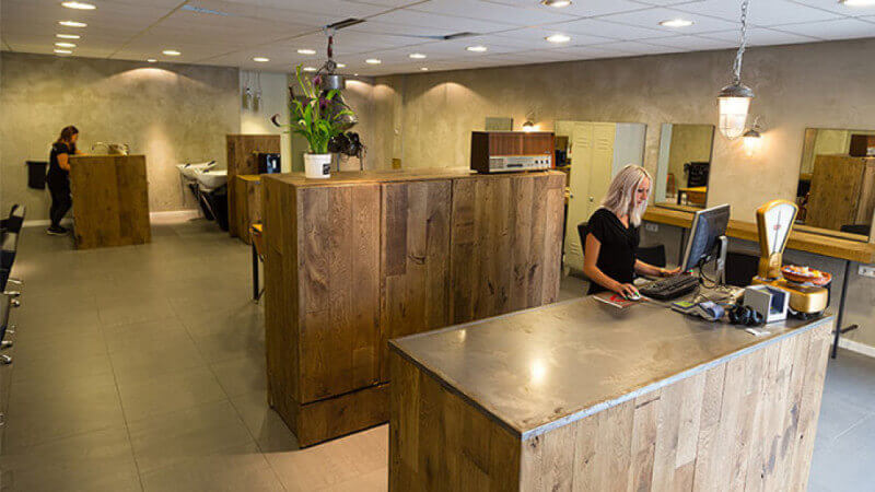 A wooden front counter at a barbershop with an associate standing behind it and the rest of the salon in the background.