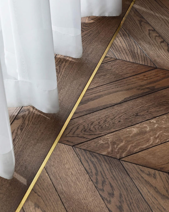 Close-up of white oak chevron parquet finished with Rubio Monocoat Oil Plus 2C hardwax oil, showcasing the wood's natural grain and warm custom tone, with a brass inlay border and sheer curtains in the background.