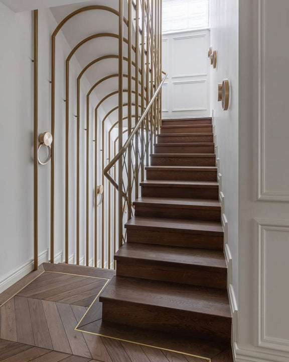 White oak staircase finished with Rubio Monocoat Oil Plus 2C hardwax oil, with a custom brass arched railing, circular brass wall sconces, and chevron parquet flooring with brass border inlays at the base.