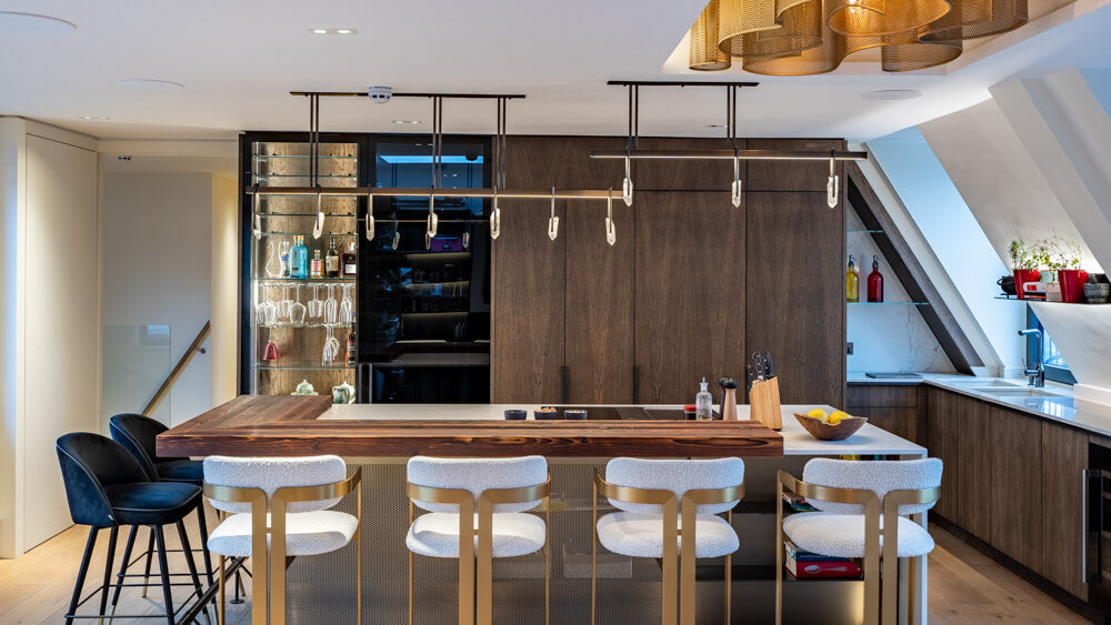 A contemporary kitchen with four white barstools and two black ones sitting at an island with a white stone countertop. A modern light fixture hangs above the island and dark oak cabinetry is in the background.