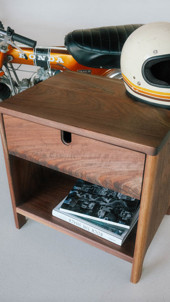 Modern walnut side table with an open bottom and drawer with an integrated finger pull on the top. A motorcycle helmet sits on top of the table, books on the lower shelf and a motorcycle in the background.
