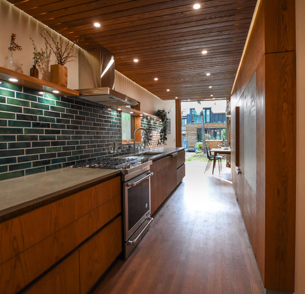 White oak cabinetry and flooring in a modern kitchen featuring a green tile backsplash.