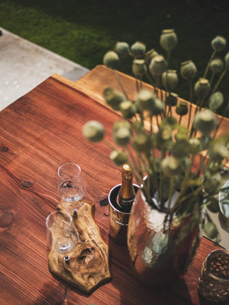 Two wine glasses, a bottle of wine and a vase with flowers are sitting on top of a redwood dining table finished with an exterior hardwax oil wood finish.
