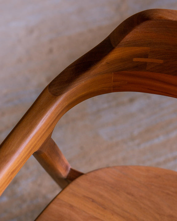 Close-up of a chair's curved backrest and armrest junction, with visible butterfly joint inlays in the wood grain, shot against a concrete floor finished with Rubio Monocoat Oil Plus 2C hardwax oil.