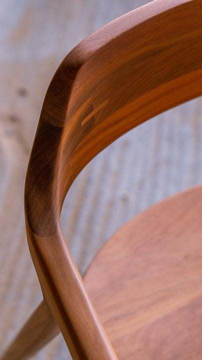 Detail of a chair's sweeping backrest curve from the side, highlighting the smooth hardwax oil wood finish and rich variation in wood tone, with a light wood-plank background.