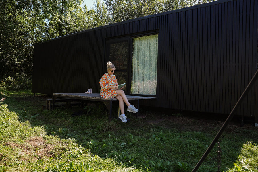 A woman is sitting on the edge of a wood deck reading a book with the black slow cabin finished with Rubio Monocoat DuroGrit is in the background.