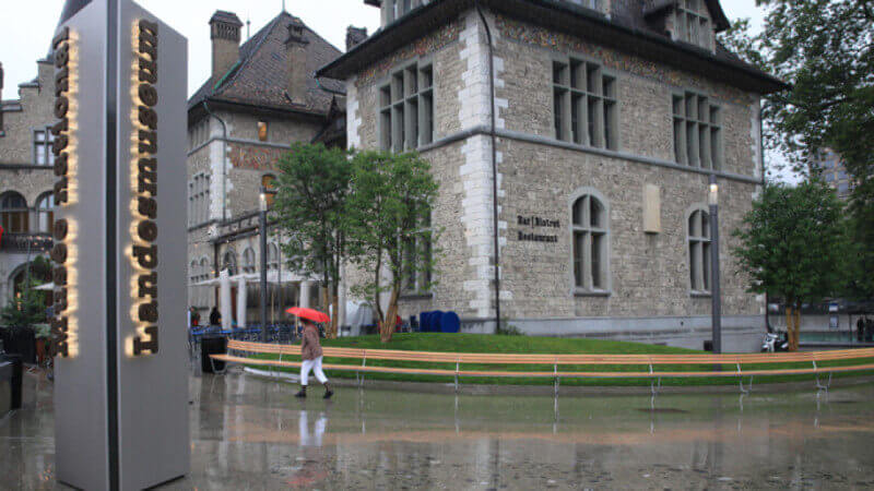 A winding wooden bench in from of the Swiss National Musuem during the rain.