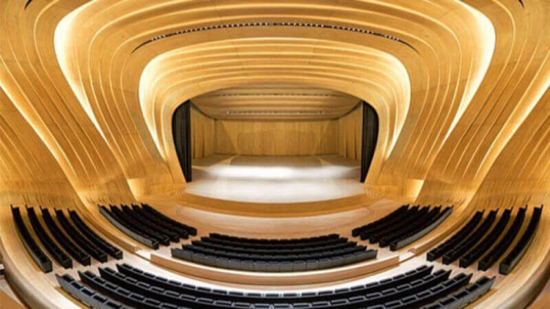 A symmetrical view of a large wood auditorium that has sweeping wood walls, recessed accent lighting, seating, and a massive wooden stage.