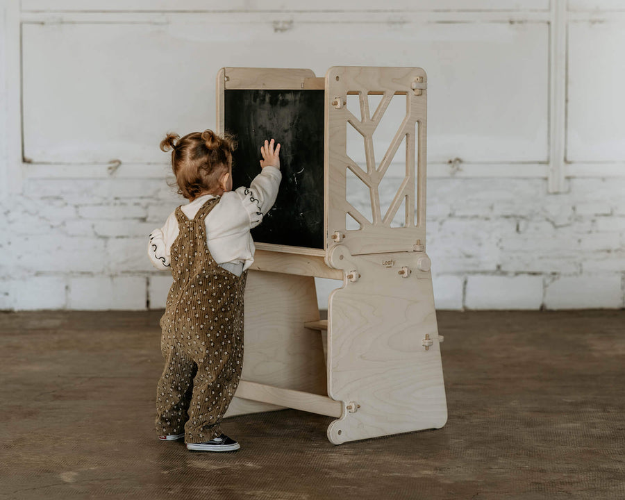 A child stands at the front side of a toddler learning tower chalk board and plays with the chalk dust.