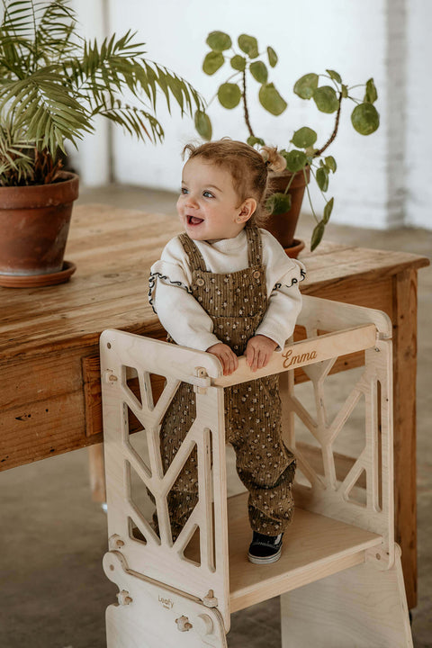 A child stand on the platform of the toddler learning tower, smiling at something off in the distance.