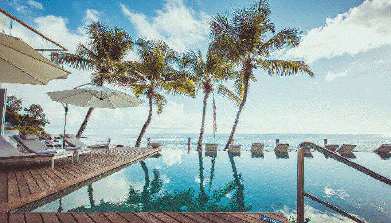 An infinity pool with a wood deck finished with an oil finish overlooking a tropical beach with palm trees.